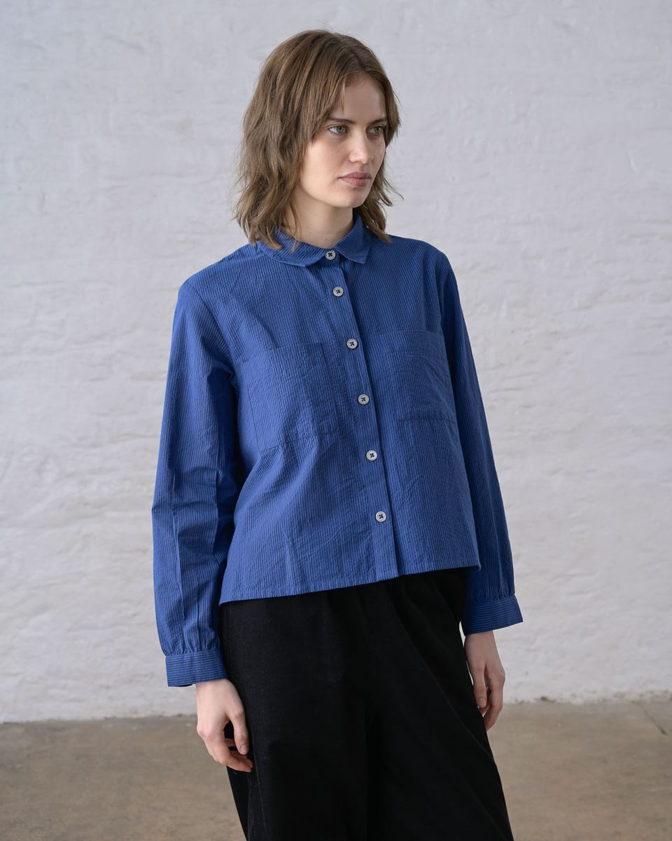Three-quarter front view of a woman wearing the BIBICO Ada Cropped Blouse in Blue Petrol Stripe, showing the rounded collar, button front with imitation shell buttons, chest patch pockets, elasticated cuffs, and boxy cropped silhouette in fine-woven blue petrol cotton stripe, styled with black wide-leg pants against a whitewashed brick wall