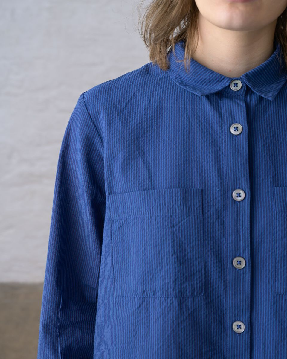 Close detail of the neckline and placket of the BIBICO Ada Cropped Blouse in Blue Petrol Stripe, showing the rounded collar, imitation shell buttons, chest patch pocket, and the fine vertical stripe texture of the blue petrol cotton fabric, against a whitewashed brick wall
