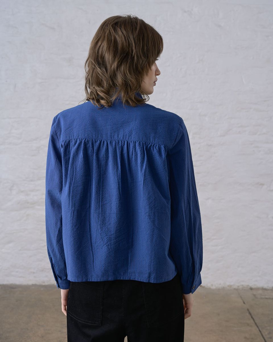 Back view of a woman wearing the BIBICO Ada Cropped Blouse in Blue Petrol Stripe, showing the gathered back yoke, full-length sleeves, and boxy cropped silhouette in fine-woven blue petrol cotton stripe, styled with black wide-leg pants against a whitewashed brick wall

