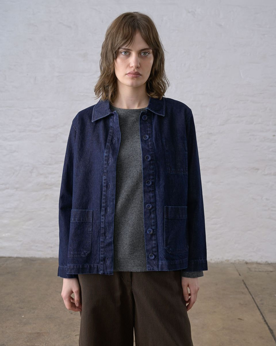 Front view of a woman wearing the BIBICO Marcia Jacket in Denim open over a gray tee, showing the pointed collar, full button front, two patch pockets, and straight boxy hem in deep indigo 11.5oz cotton denim, styled with brown wide-leg corduroy pants against a whitewashed brick wall
