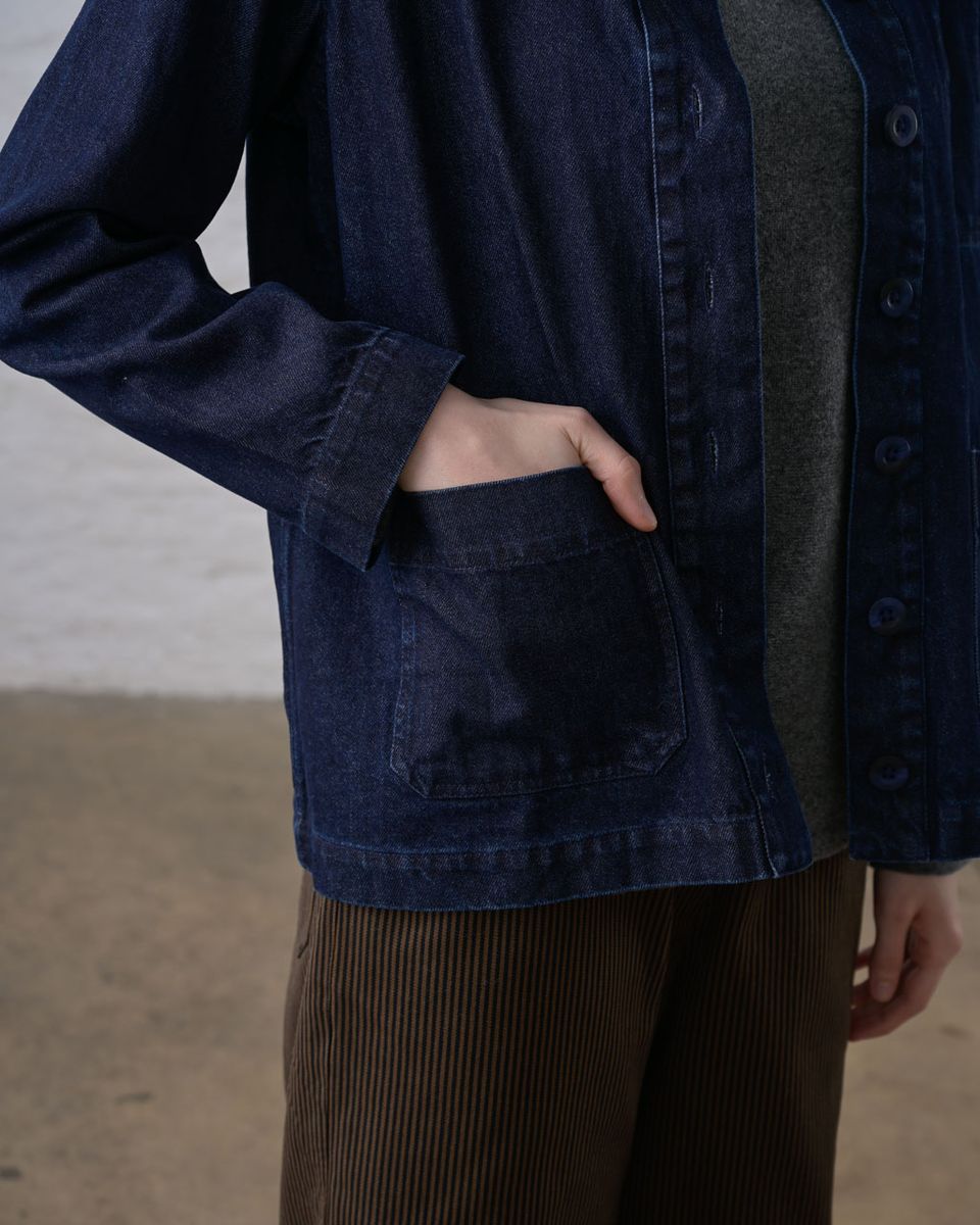 Close detail view of the hip area of the BIBICO Marcia Jacket in Denim, showing a hand inside the deep patch pocket, topstitching, button placket, and the weight and texture of the indigo 11.5oz cotton denim, with a gray tee and brown corduroy pants visible underneath
