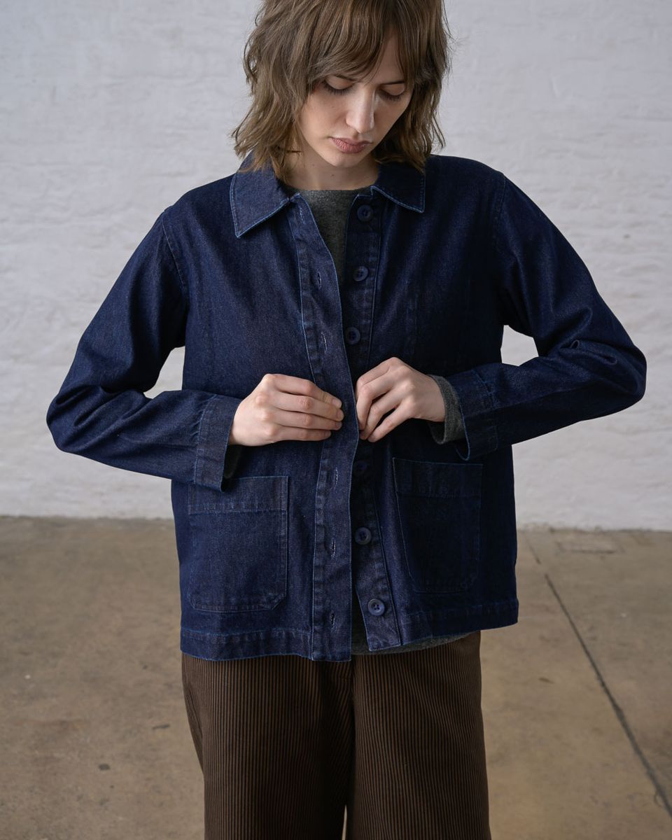 Three-quarter front view of a woman buttoning the BIBICO Marcia Jacket in Denim, showing the pointed collar, full button placket, two hip patch pockets, and topstitching detail in deep indigo 11.5oz cotton denim, styled with a gray tee and brown wide-leg corduroy pants against a whitewashed brick wall
