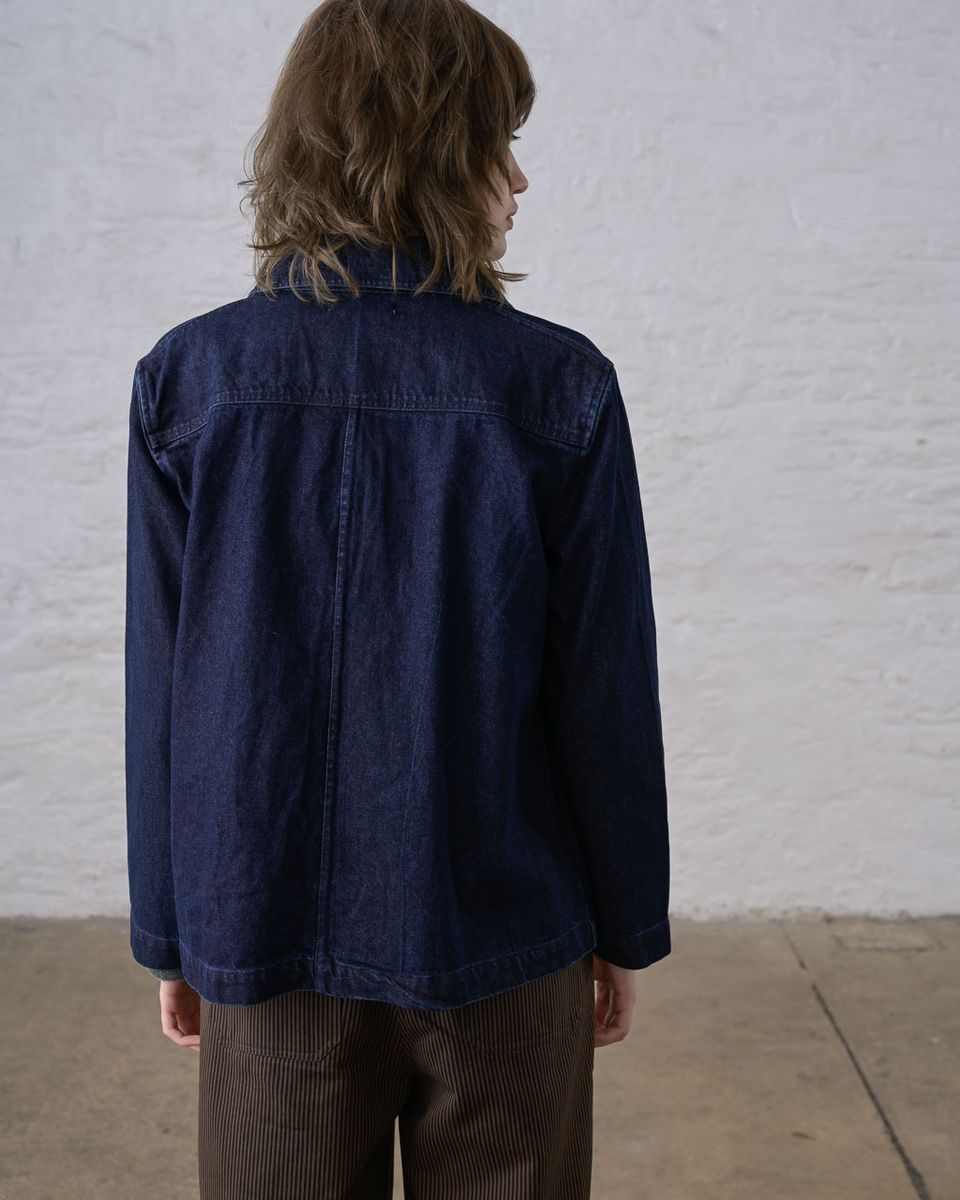 Back view of a woman wearing the BIBICO Marcia Jacket in Denim, showing the center back seam, structured yoke, full-length sleeves, and straight boxy hem in deep indigo 11.5oz cotton denim, styled with brown wide-leg corduroy pants against a whitewashed brick wall
