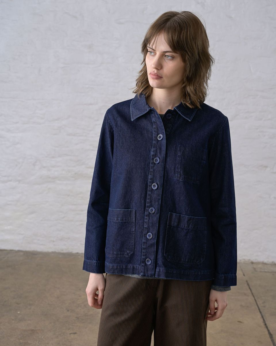 Full-length front view of a woman wearing the BIBICO Marcia Jacket in Denim buttoned closed, showing the pointed collar, button placket, two hip patch pockets, and high-hip length silhouette in deep indigo 11.5oz cotton denim, styled with brown wide-leg corduroy pants against a whitewashed brick wall in natural light
