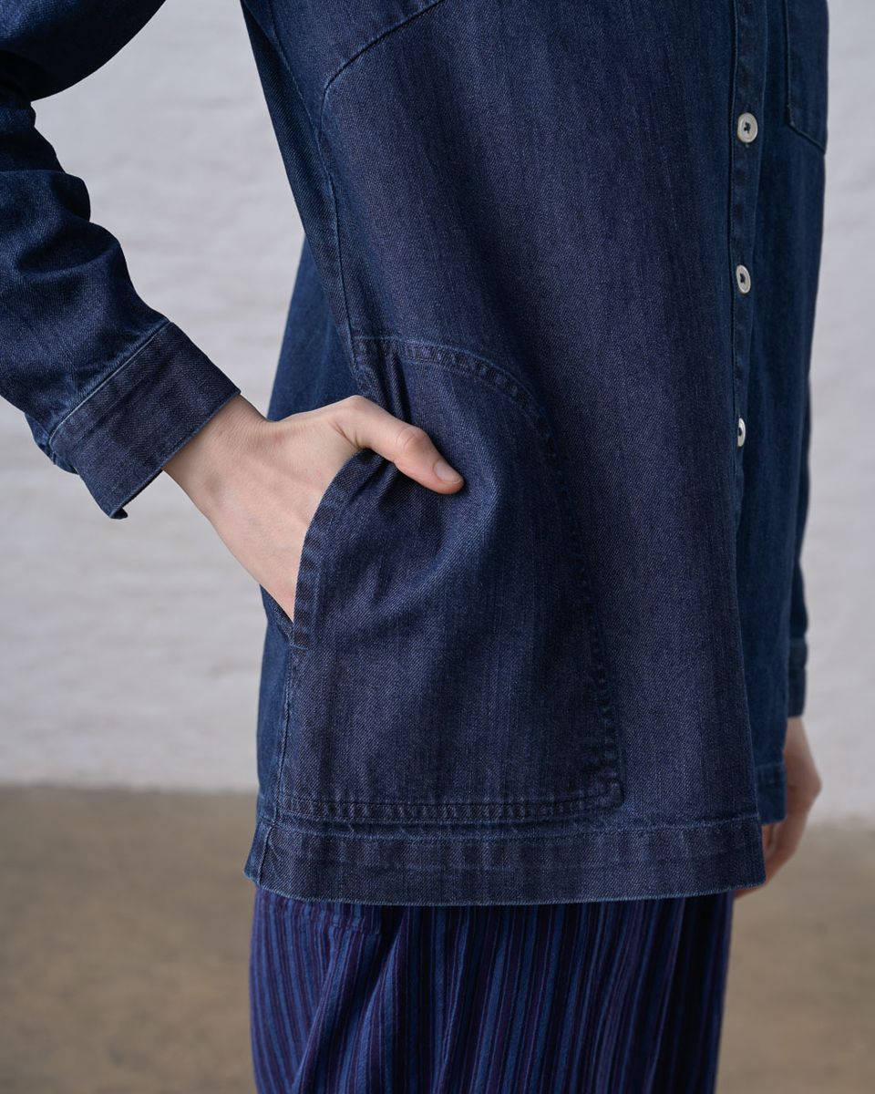 Close side detail of the BIBICO Miriam Tunic in Denim, showing a hand in the large side hip pocket, button placket, buttoned cuff, and the weight and drape of the deep indigo 9oz cotton denim, styled over a blue pleated skirt against a whitewashed brick wall
