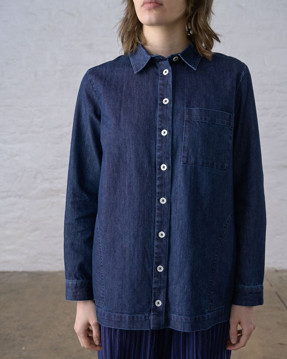 Cropped front view of a woman wearing the BIBICO Miriam Tunic in Denim buttoned closed, showing the pointed collar, full white button placket, single chest patch pocket, and relaxed fit in deep indigo 9oz cotton denim, styled with a blue pleated skirt against a whitewashed brick wall
