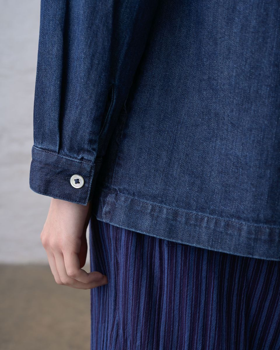 Close detail of the lower side of the BIBICO Miriam Tunic in Denim, showing a hand reaching into the roomy side hip pocket, the white buttoned cuff, curved shirttail hem, and deep indigo 9oz cotton denim fabric, styled over a blue pleated skirt
