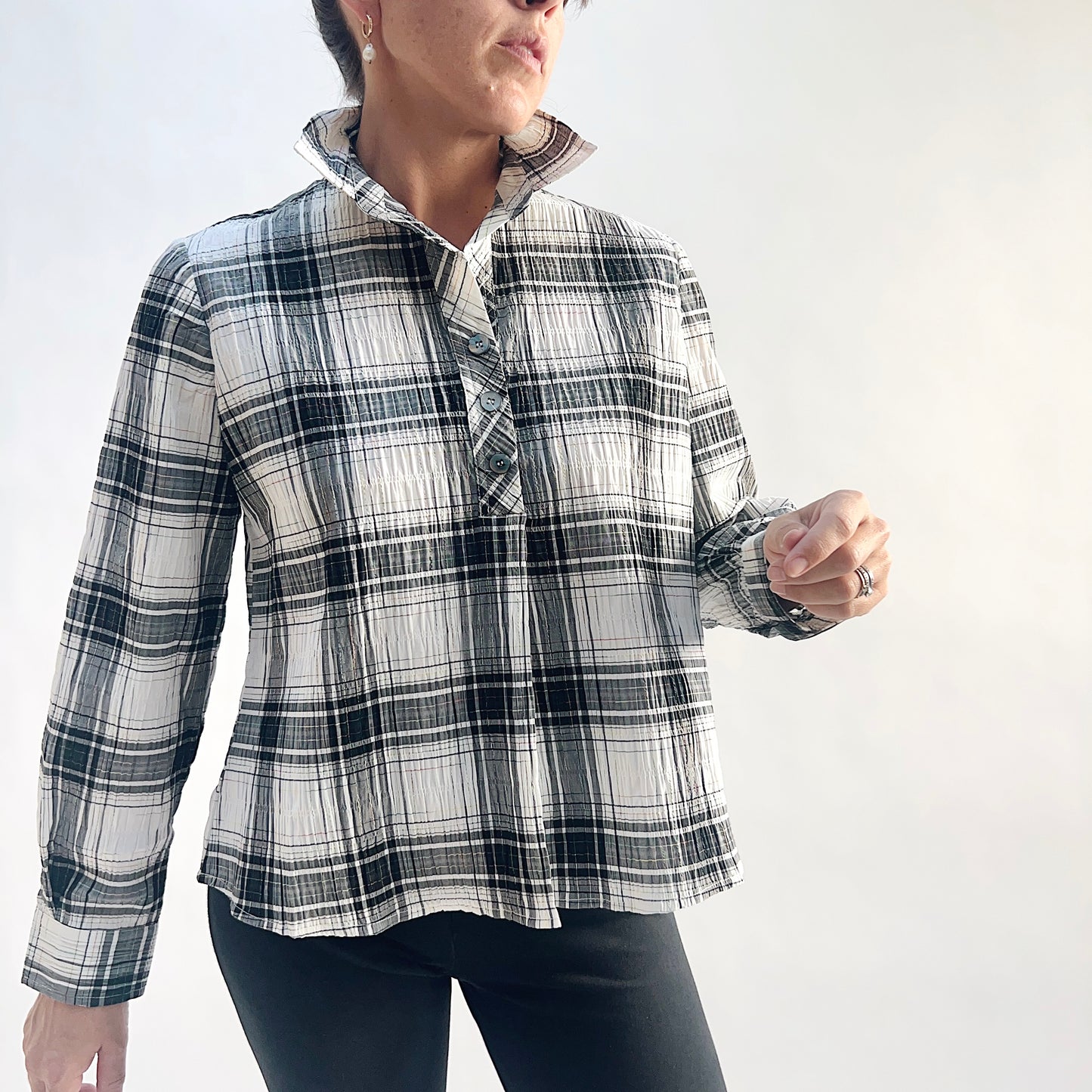 Close front view of model wearing a white and charcoal windowpane plaid crinkle pullover shirt with point collar open at the neck, showing the half placket with matte buttons and long sleeves, styled with black slim pants against a white background.
