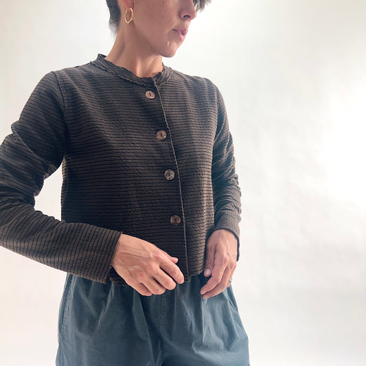 Three-quarter front view of model wearing a chocolate and black fine tonal stripe cropped cardigan with asymmetrical button closure and long sleeves, hands at hem, showing the band neckline and crinkle-texture stripe detail, styled with teal wide-leg pants against a white background.
