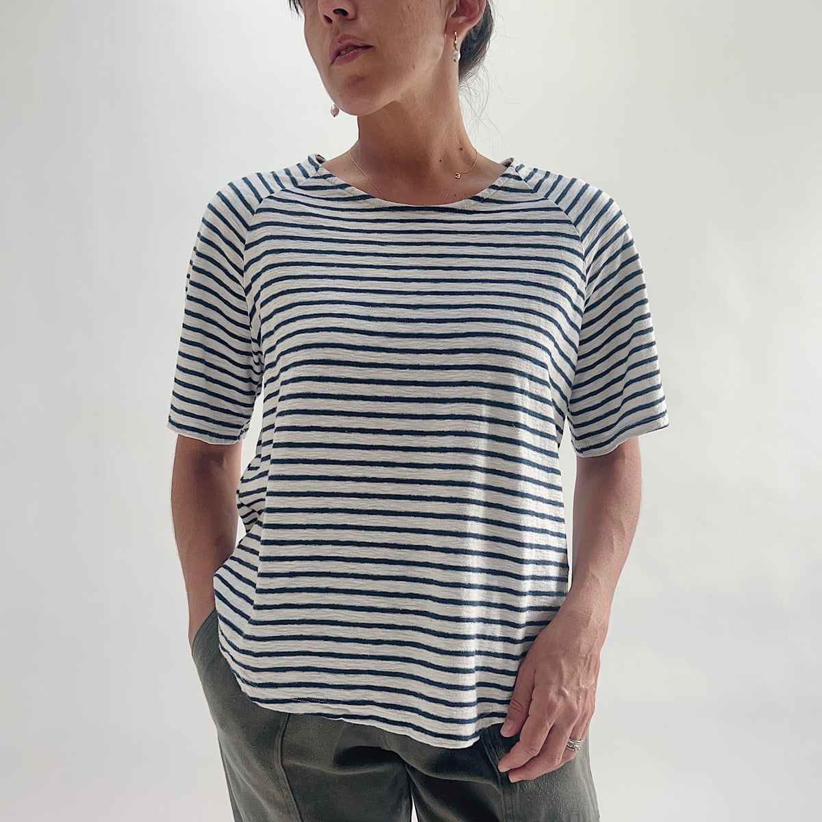 Close front view of model wearing a navy and white striped boxy raglan tee with hands on hips, showing the boat neckline and short raglan sleeves.
