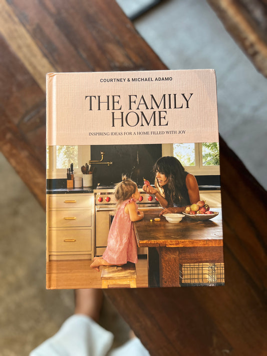 The Family Home by Courtney and Michael Adamo, photographed on a wooden surface, with a woman and child in a kitchen on the cover.
