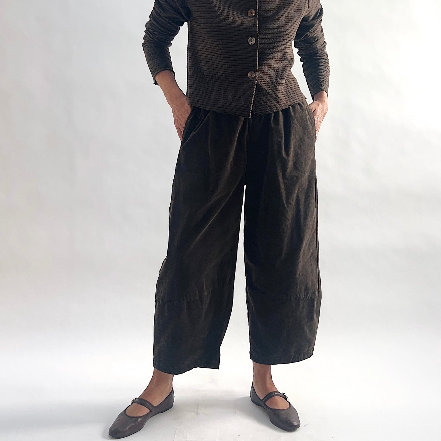 Full front view of model wearing chocolate mini cord lantern pants with hands in side pockets, showing the gathered elastic waist, full lantern volume, and tapered ankle, styled with the matching striped cropped cardigan and gray mary jane flats against a white background.
