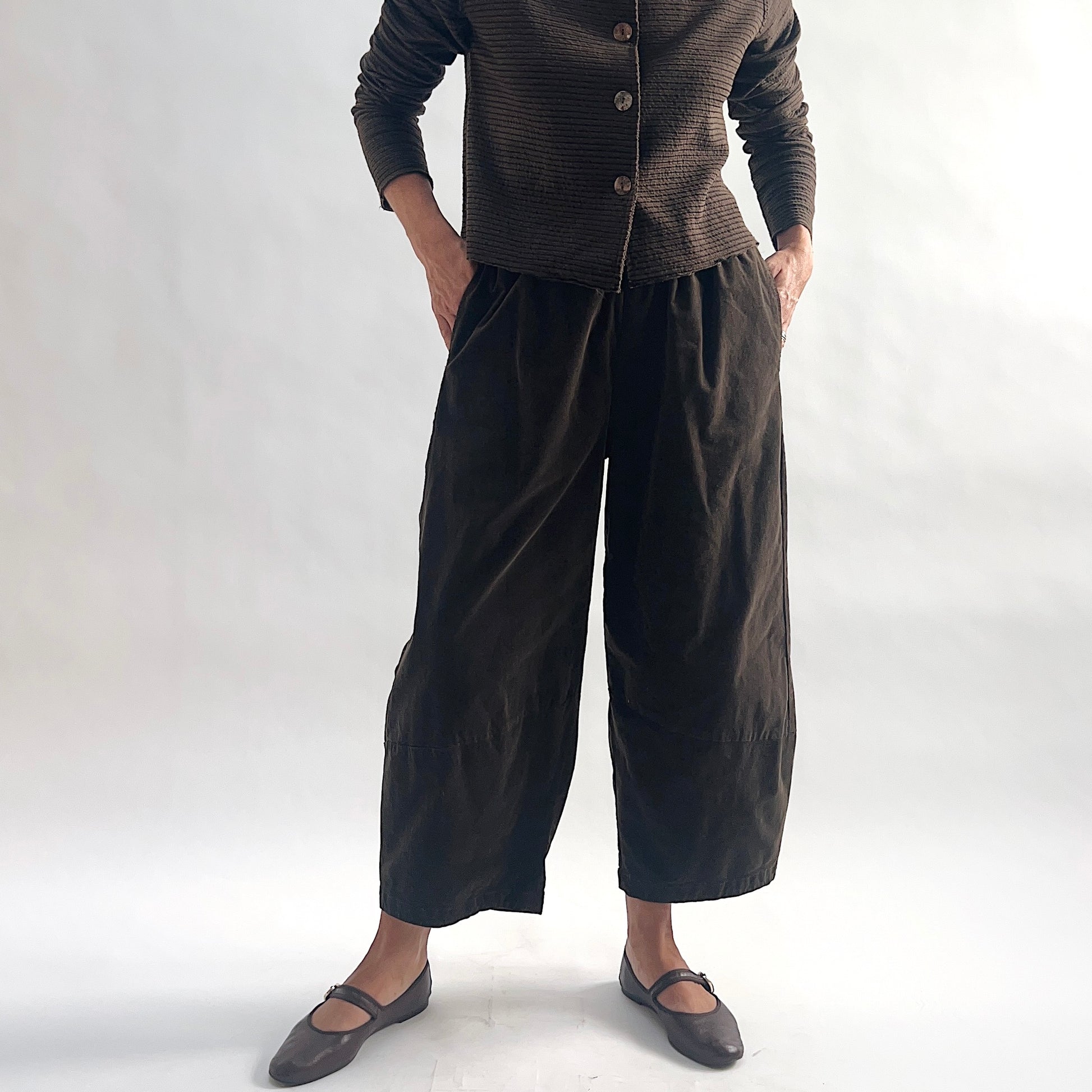 Full front view of model wearing chocolate mini cord lantern pants with hands in side pockets, showing the gathered elastic waist, full lantern volume, and tapered ankle, styled with the matching striped cropped cardigan and gray mary jane flats against a white background.
