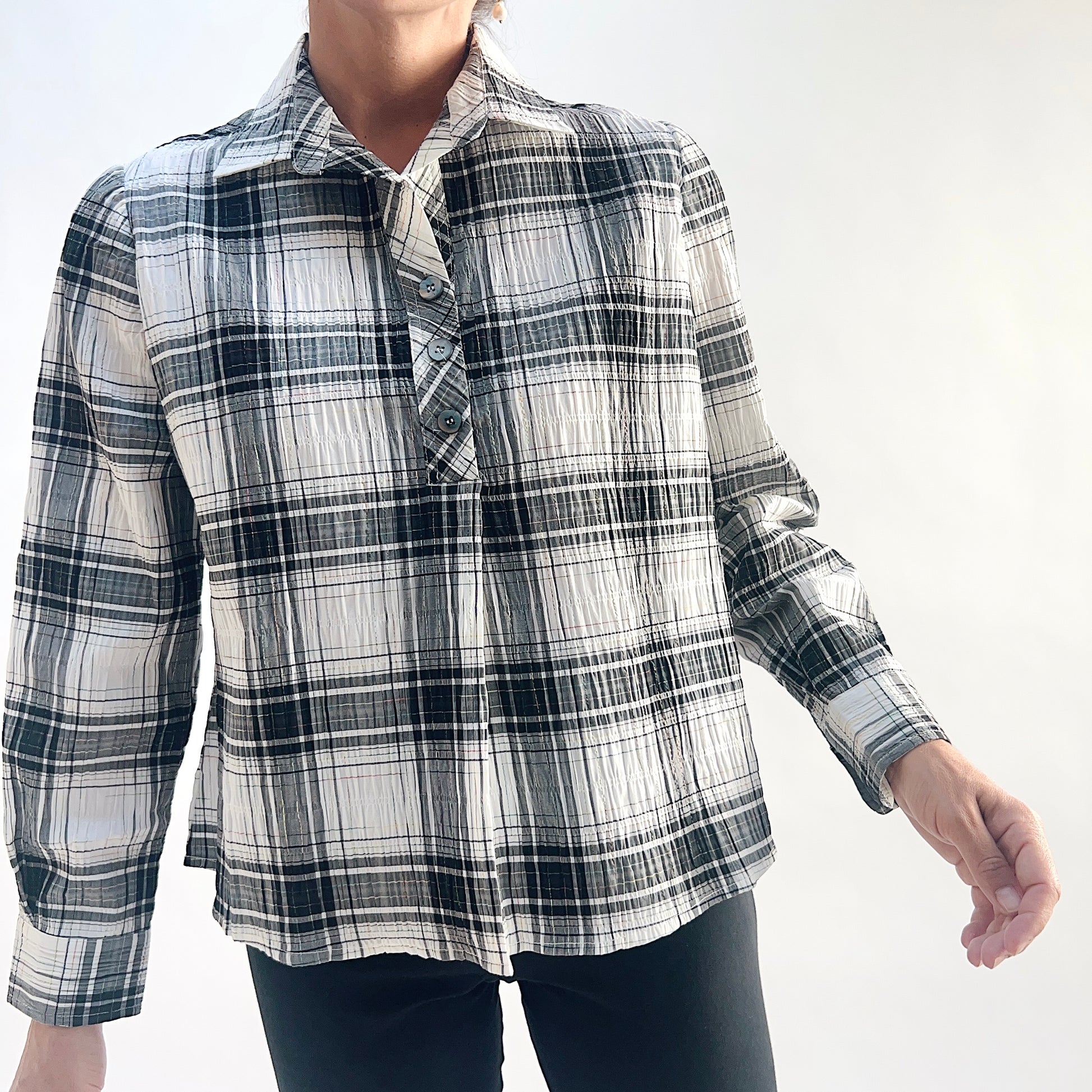 Three-quarter front view of model wearing a white and charcoal windowpane plaid crinkle pullover shirt, arm extended, showing the point collar open at the neck, half placket with matte buttons, and buttoned cuffs, styled with black slim pants against a white background.
