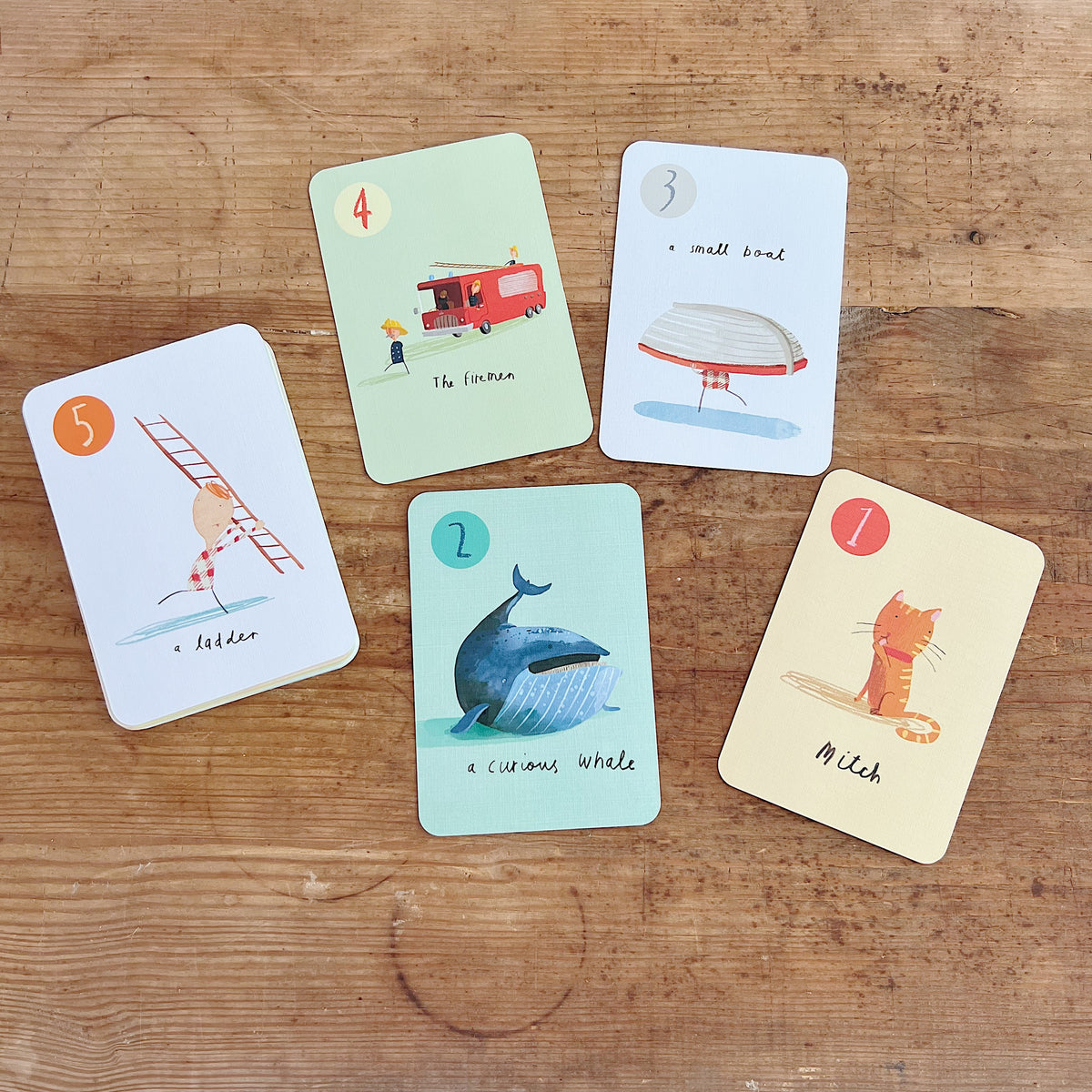 Five Go Fish cards by Oliver Jeffers spread on rustic wooden surface, showing illustrated characters including a curious whale, a cat named Mitch, a small boat, a ladder, and the firemen
