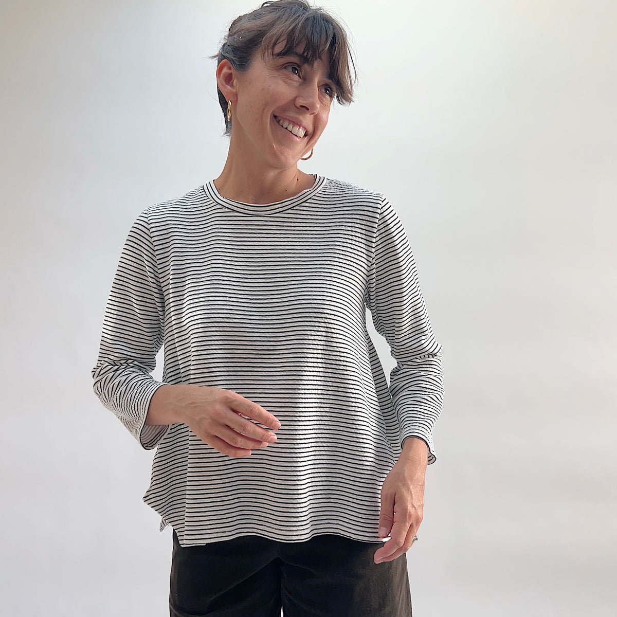 Cut Loose Striped Side Vent Crew in Laundered, white and black fine-stripe crewneck top with relaxed boxy fit, model smiling and looking to the side, paired with dark wide-leg trousers — front view
