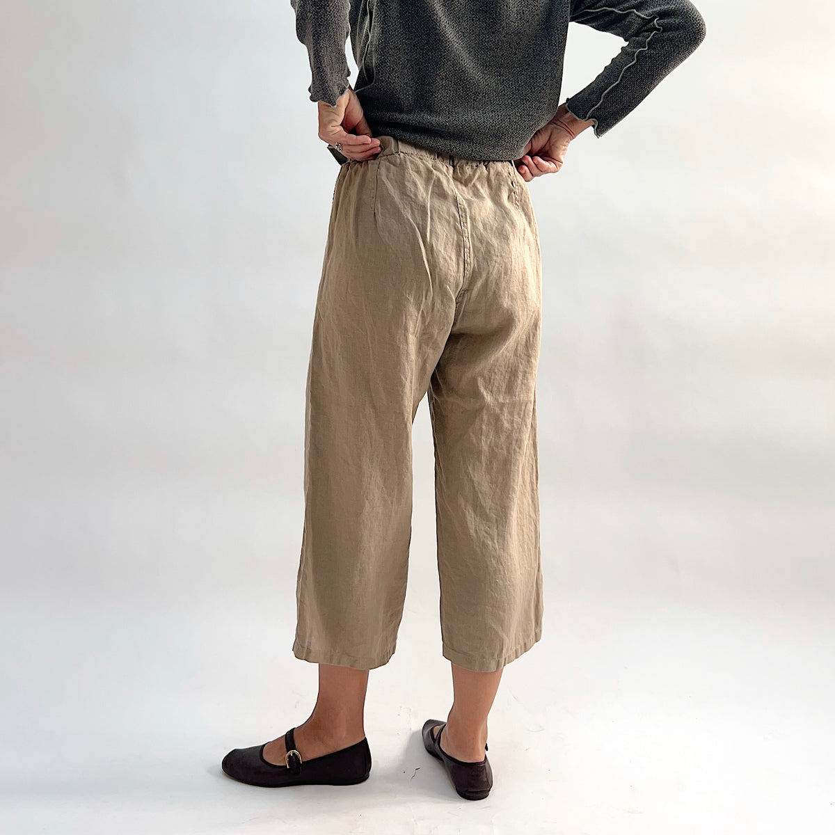 Cut Loose Hanky Linen Easy Crop Flood Pant in Suede, sandy tan linen wide-leg crop pant with hands on hips, styled with a grey button-front cardigan and brown Mary Jane flats — back view showing elastic waistband and straight hem
