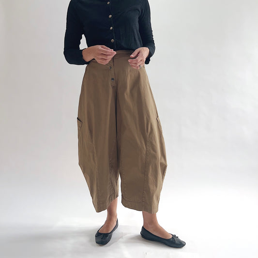Close front view of model wearing Eleven Stitch winter olive snap front pant, showing snap front waistband, wide balloon leg, large side patch pockets, gathered elastic ankle hem, and warm olive tan color, styled with black button front top and black ballet flats