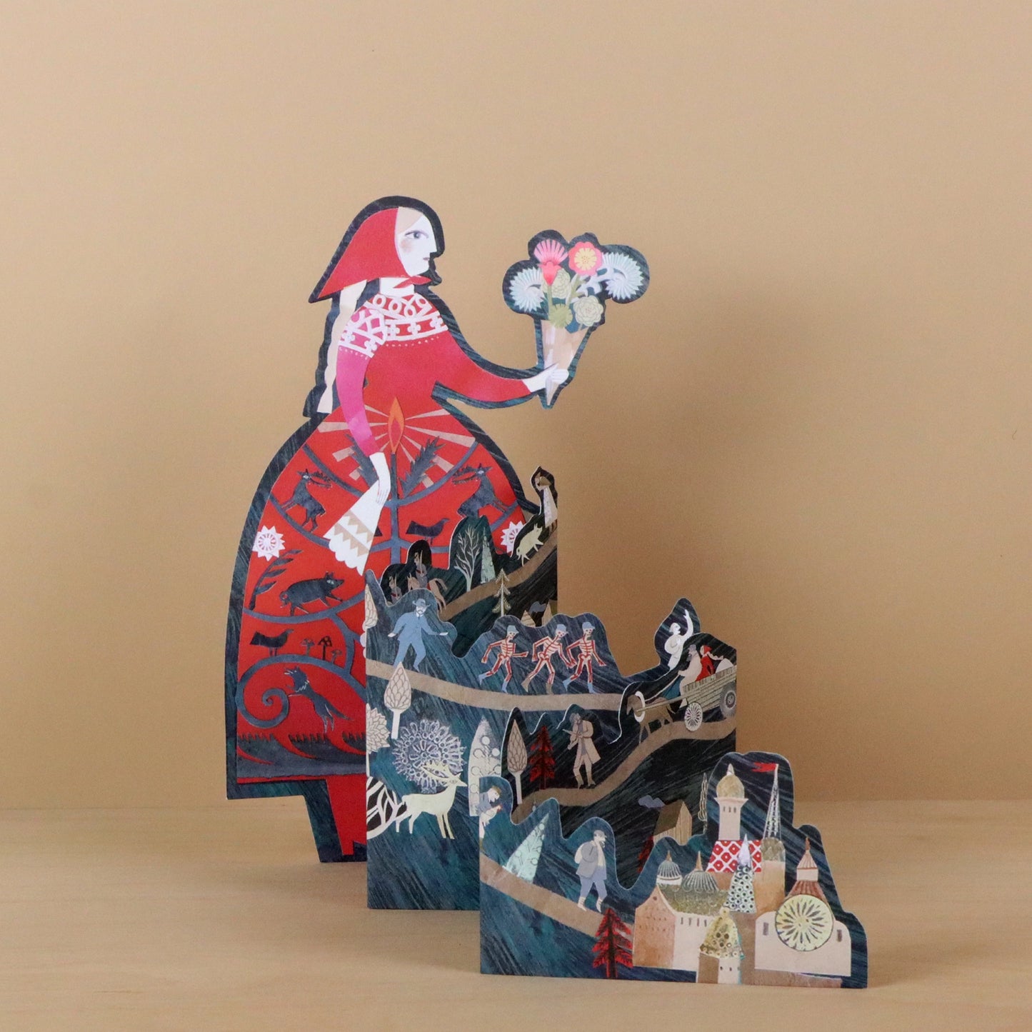 Storyteller die-cut concertina card by Sarah Young, angled view showing a tall woman in a red folkloric dress holding flowers, with layered concertina panels below depicting fairy tale scenes including figures, a sleigh, and a village, against a warm beige background