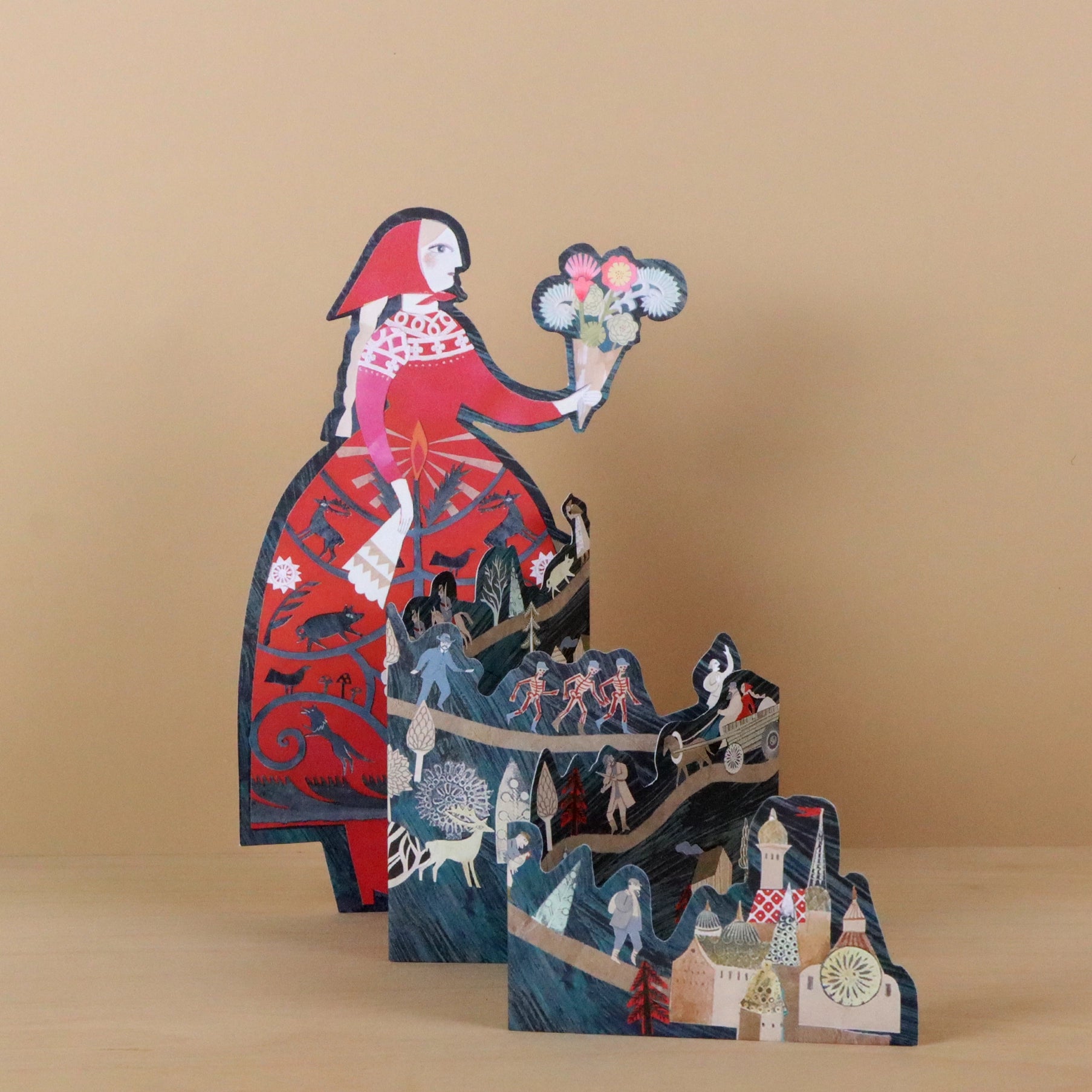 Storyteller die-cut concertina card by Sarah Young, angled view showing a tall woman in a red folkloric dress holding flowers, with layered concertina panels below depicting fairy tale scenes including figures, a sleigh, and a village, against a warm beige background