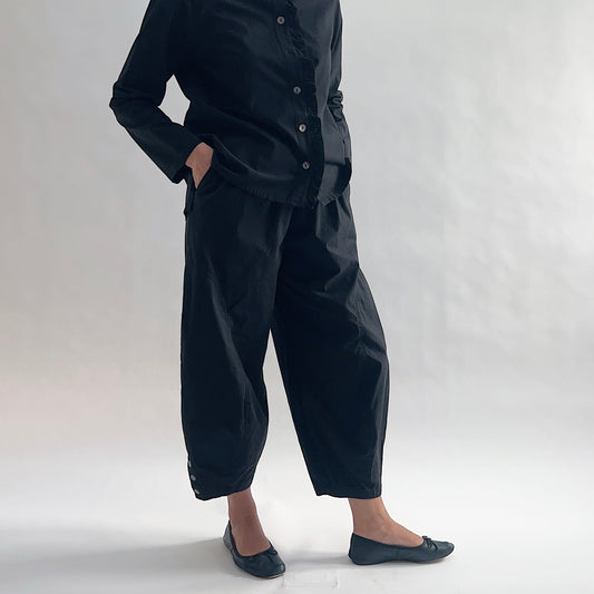 Three-quarter side view of model wearing Eleven Stitch black snap doreen pant, showing elastic waistband, wide balloon leg tapering to snap hem at ankle, side seam detailing, snap buttons at ankle, and black color, styled with matching black blossom collar shirt and black ballet flats