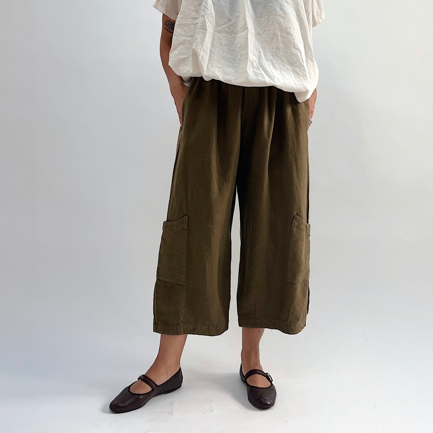Model wearing the Bryn Walker Heavy Linen Casbah Pant in Partridge, full front view highlighting elastic waistband, two patch pockets, cropped wide-leg hem, and olive linen texture, with dark Mary Jane flats
