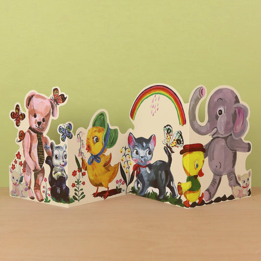 Plushie Party die-cut concertina card by Nathalie Lété, partially open showing a pink teddy bear with a tie and a skunk alongside a black cat, yellow chick, and elephant, with butterflies and a rainbow, against a green background