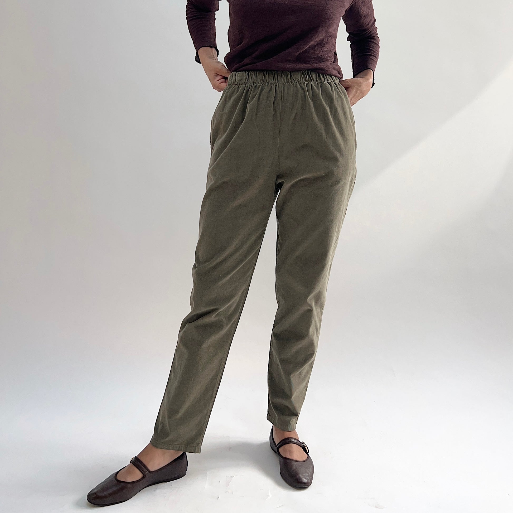 Model with hands in pockets wearing the Bryn Walker Corduroy Boslow Pants in Jasper, full front view highlighting fully elastic waistband, side pockets, relaxed tapered silhouette, and olive jasper pinwheel corduroy, styled with burgundy Double Cotton Tee and brown Mary Jane flats

