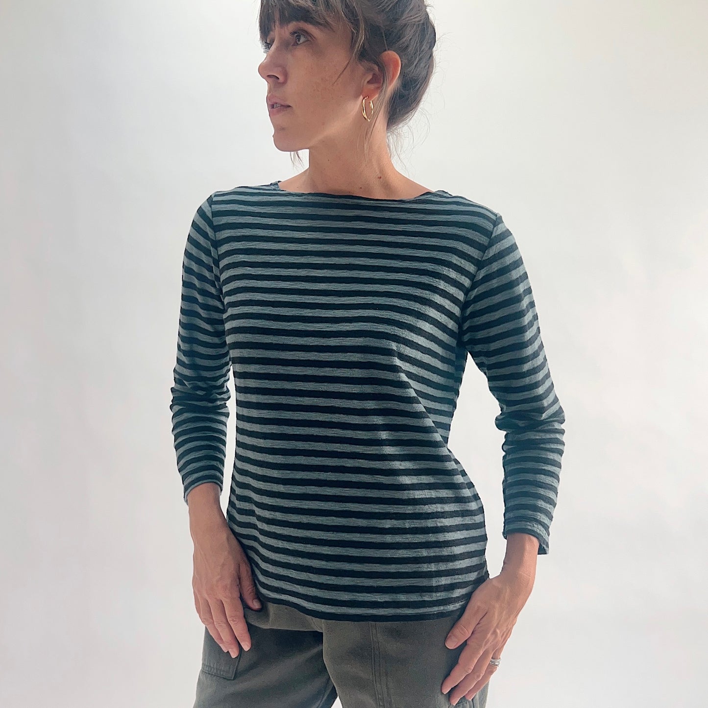Front view of model wearing an iron stripe 3/4 sleeve boatneck tee in a cotton-linen blend, looking over her shoulder, styled with olive green pants against a white background.
