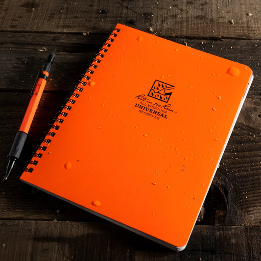 Bright orange spiral-bound notebook with black coil and a matching orange pen placed beside it on a dark wooden surface. The notebook has a printed logo and the word UNIVERSAL on the cover, and water droplets are visible on the surface.