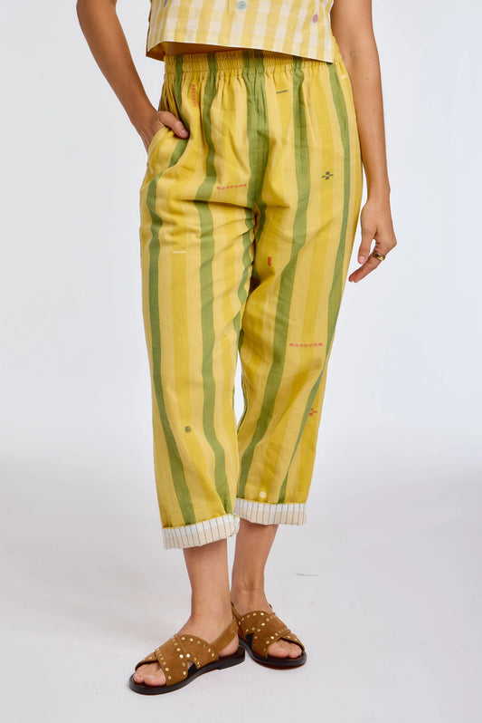 Close-up front view of DVAA Celeste Pant in Goldenrod against white studio backdrop showing goldenrod yellow and sage green wide vertical stripe handwoven cotton with scattered small ikat motifs, elasticated drawstring waistband, hand in side pocket, cropped tapered leg with contrast white pinstripe cuff turn-up, styled with tan gold-studded slide sandals
