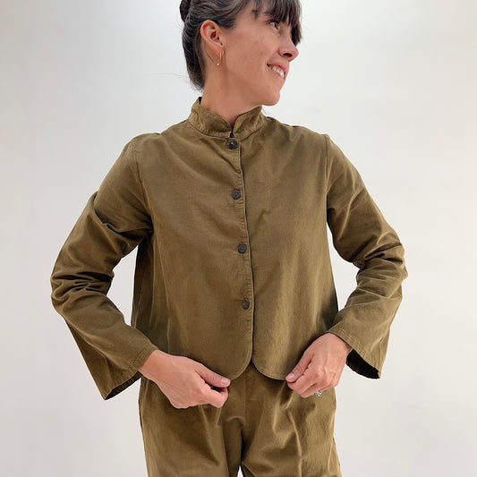 Model smiling and wearing the Bryn Walker Corduroy Standing Collar Crop Jacket in Partridge, front view showing ruffled standing collar, shell button closure, and olive pinwheel corduroy, paired with matching Pasha Pants
