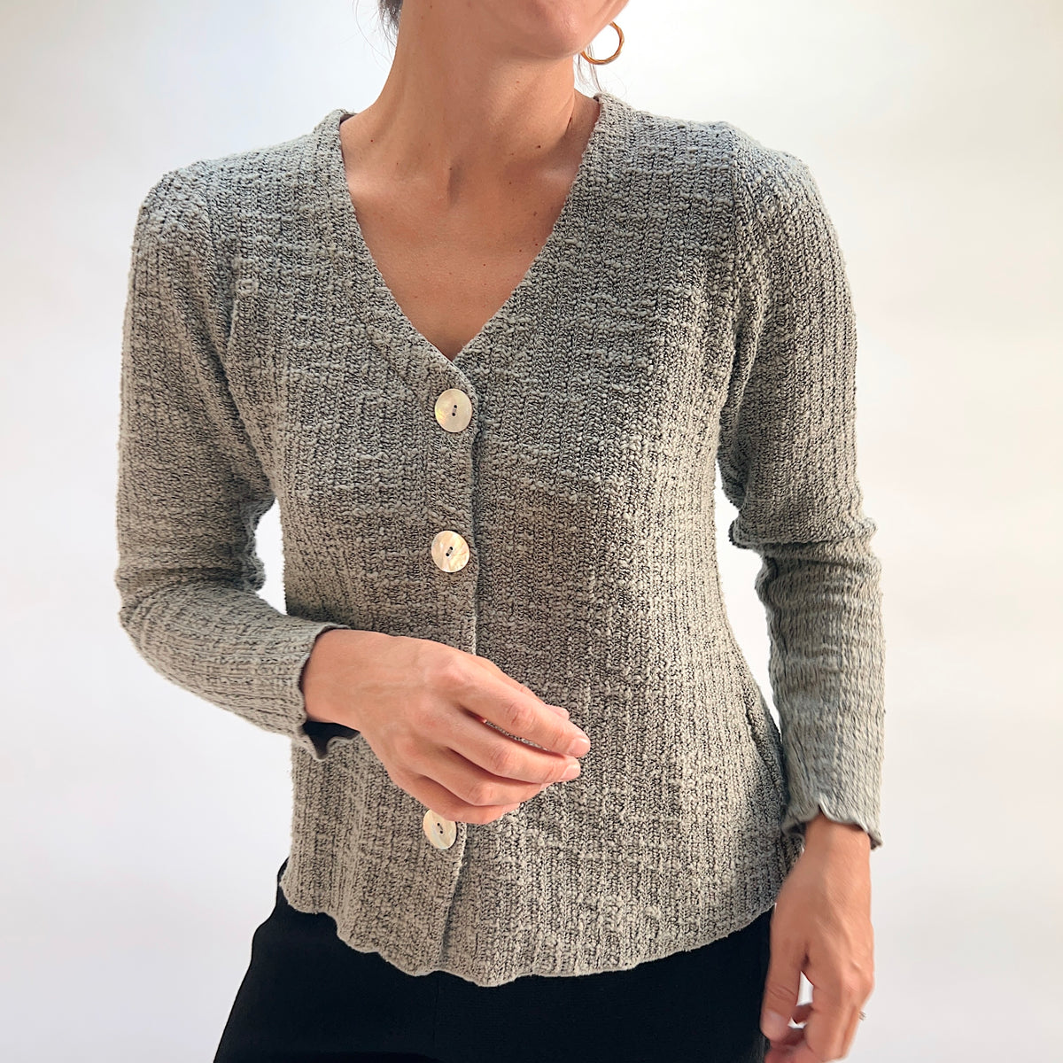 Model wearing the Cut Loose Puff Sleeve Cardigan in Squirrel, smiling three-quarter front view, showing V-neckline, natural shell button placket, gently puffed shoulders, and scalloped hem of warm grey-taupe marled knit cardigan, styled with black pants
