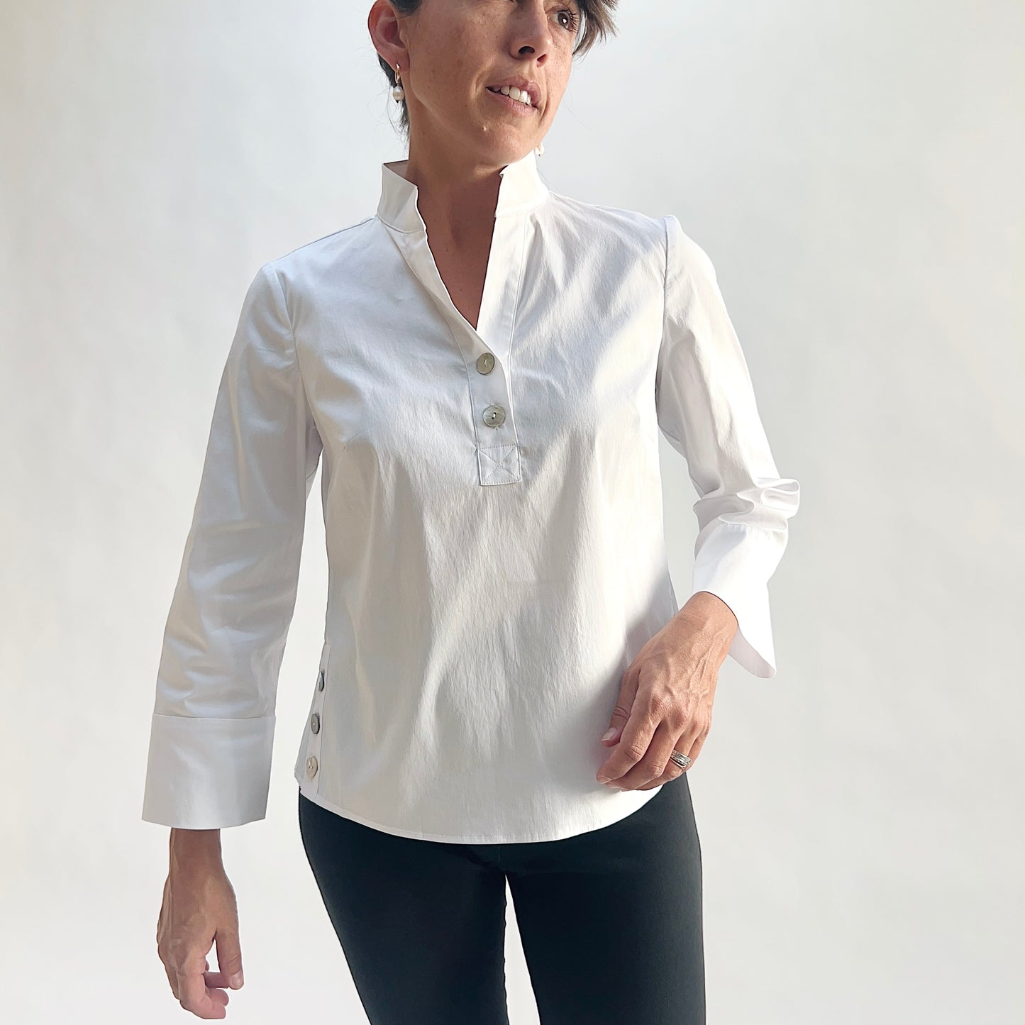Front view of model wearing a white slubby cotton pullover shirt with a standing band collar, V-notch, half placket with oversized shell buttons, and 3/4 sleeves with wide cuffs, showing the side hem button vents and curved shirttail, styled with black slim pants against a white background.
