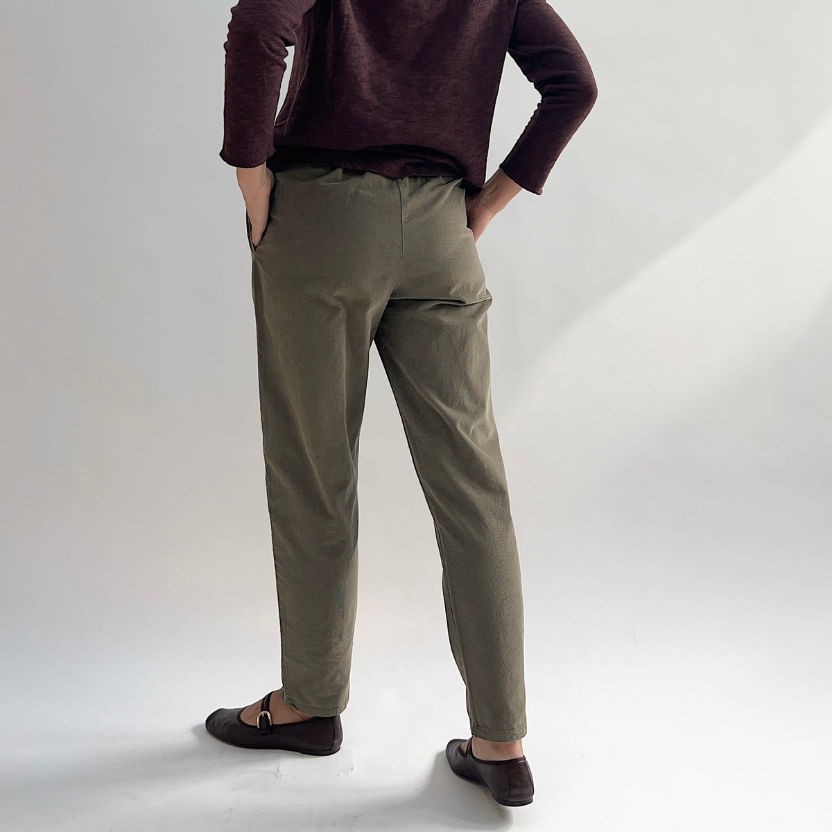 Back view of model wearing the Bryn Walker Corduroy Boslow Pants in Jasper, showing elastic waistband, relaxed tapered leg, ankle length, and olive jasper pinwheel corduroy, styled with burgundy Double Cotton Tee and brown Mary Jane flats
