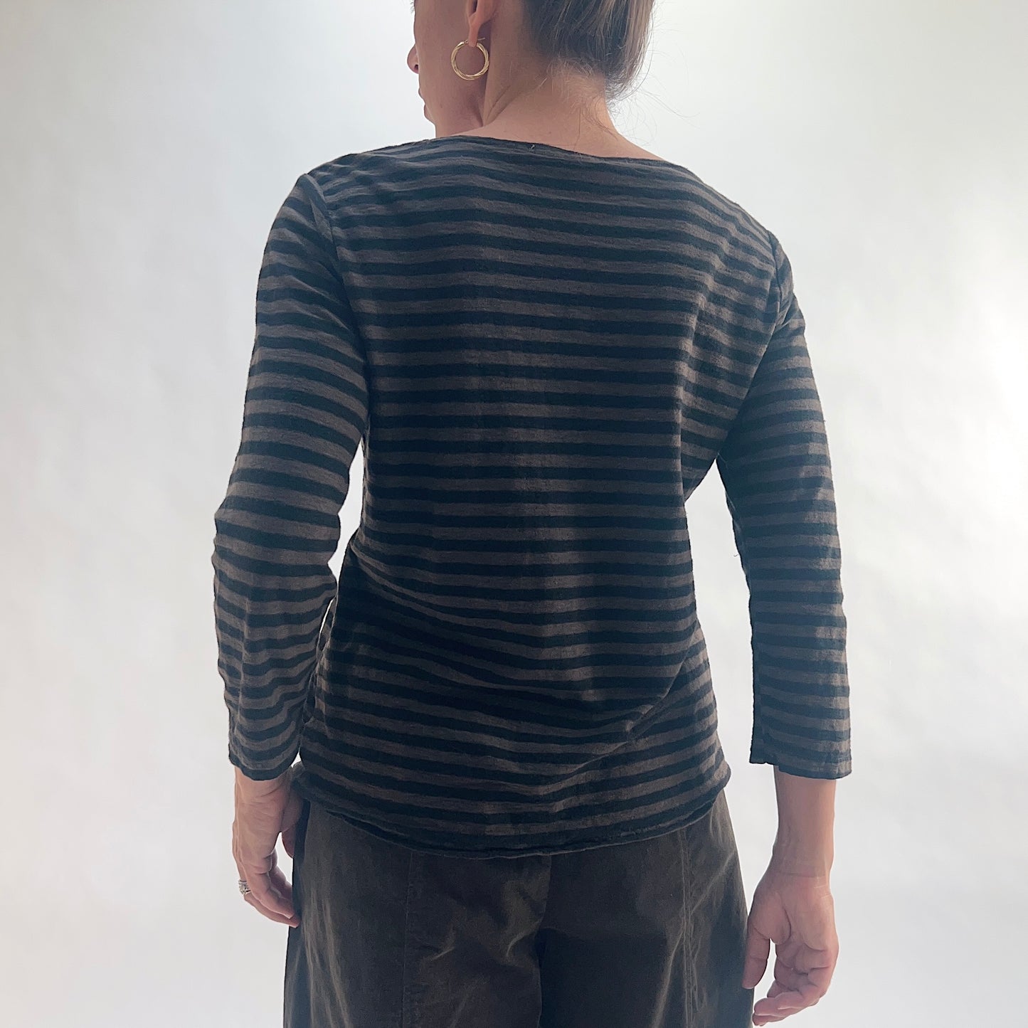 Back view of model wearing a chocolate and black horizontal stripe boatneck tee with 3/4 sleeves, showing the wide boatneck and curved hem, styled with dark olive pants against a white background.
