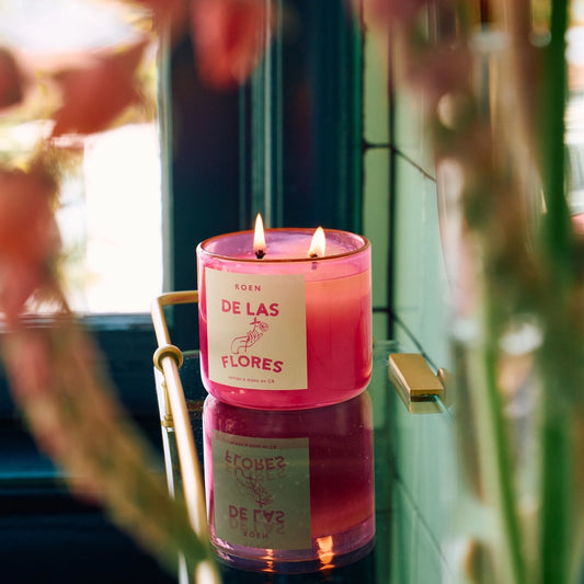A pink jar candle with multiple wicks burning inside, placed on a surface with flowers in the background.