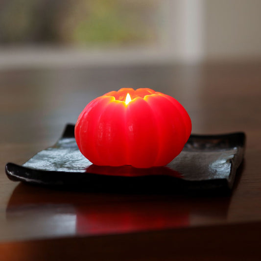 Big Dipper Wax Works Small Beeswax Pumpkin Candle in dark orange, lit and glowing on a black ceramic tray on a wooden tabletop, showing a squat ribbed pumpkin shape with a burning flame at the top and warm orange light emanating from inside, against a softly blurred indoor background
