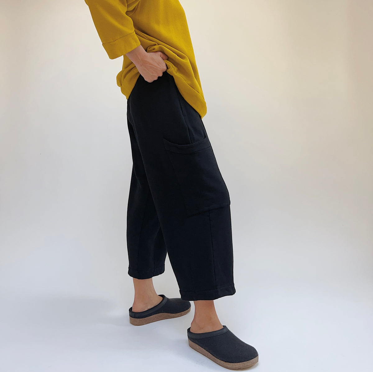 Side lower-body view of a woman wearing the Bryn Walker Pasha Fleece Pant in Black, showing the elastic waistband, large thigh patch pocket, wide straight leg silhouette, and folded ankle hem in black organic cotton and bamboo fleece, styled with a mustard yellow sweatshirt and dark slide clogs against a white background
