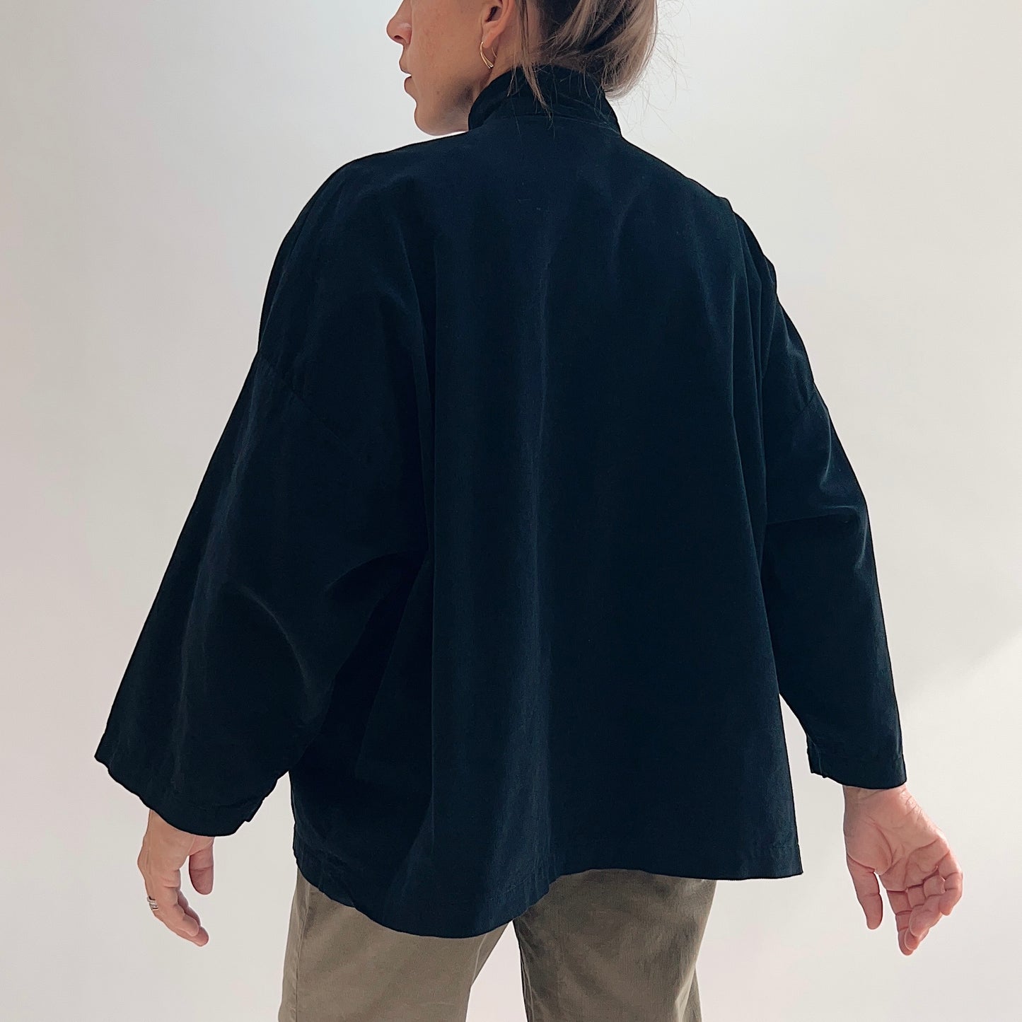 Back view of model wearing the Bryn Walker Corduroy Danuta Jacket in Black, showing standing collar, wide dolman sleeves, curved longer hem, and black pinwheel corduroy drape, styled with olive pants

