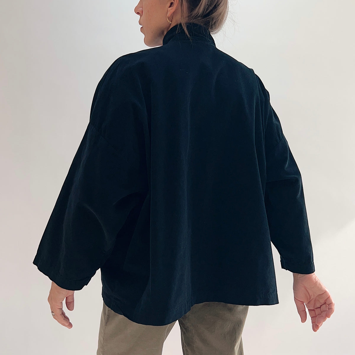 Back view of model wearing the Bryn Walker Corduroy Danuta Jacket in Black, showing standing collar, wide dolman sleeves, curved longer hem, and black pinwheel corduroy drape, styled with olive pants
