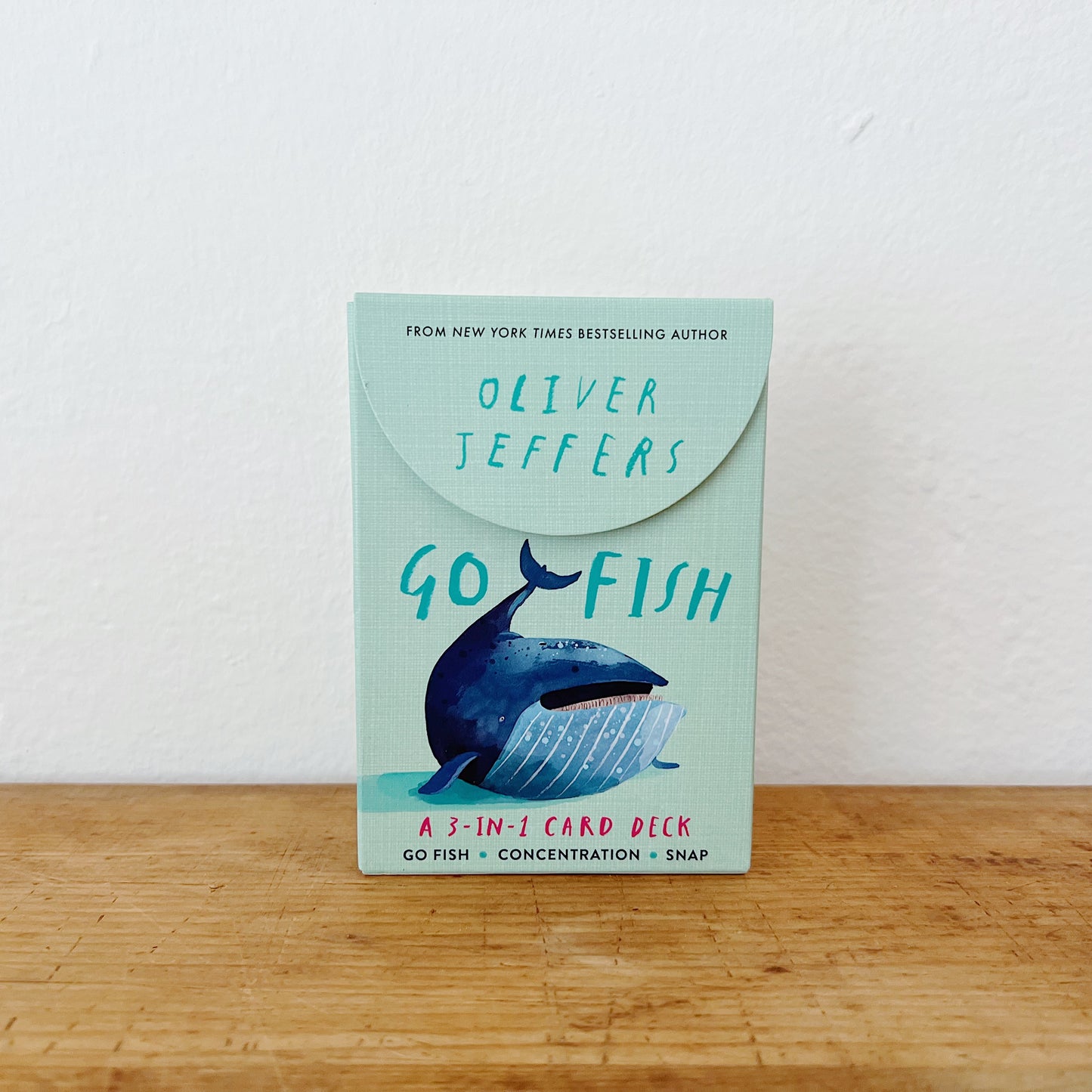 Go Fish: A 3-in-1 Card Deck by Oliver Jeffers, mint green flip-top box with illustrated whale, standing upright on wooden surface against white background
