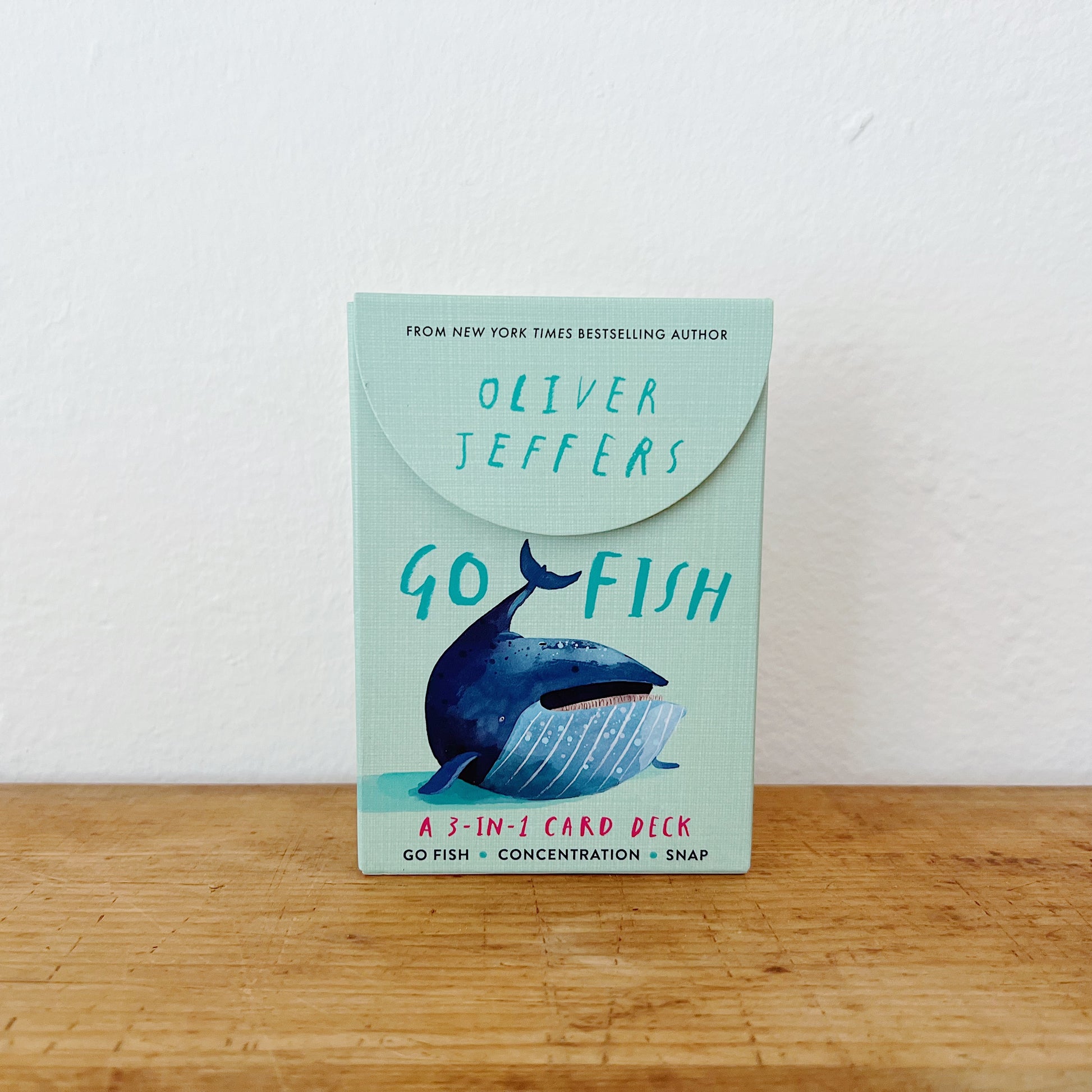 Go Fish: A 3-in-1 Card Deck by Oliver Jeffers, mint green flip-top box with illustrated whale, standing upright on wooden surface against white background
