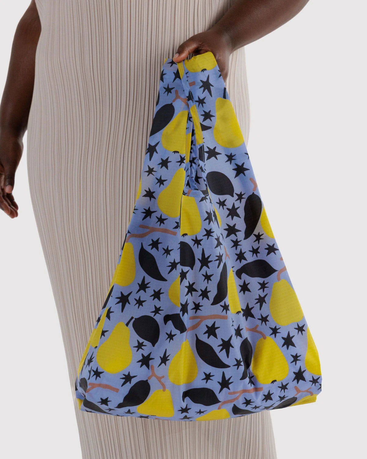 BAGGU Standard Baggu in Golden Pears, held at a model’s side, showing yellow pears, black leaves, and stars on light blue fabric.
