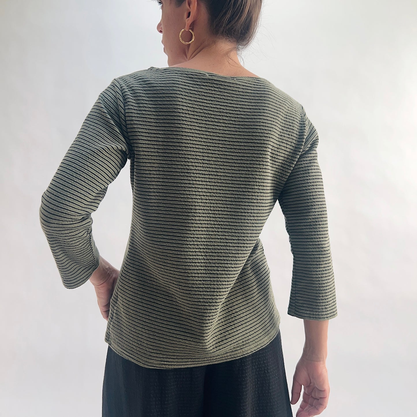 Back view of model wearing a kelp green tonal textured stripe boatneck tee with 3/4 sleeves, hand on hip, showing the wide boatneck and curved hem with crinkle-texture fabric detail, styled with a black skirt.
