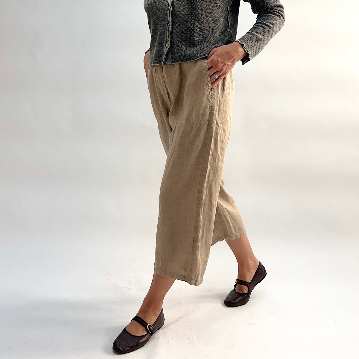 Cut Loose Hanky Linen Easy Crop Flood Pant in Suede, sandy tan linen wide-leg crop pant with hands in pocket, styled with a grey button-front cardigan and brown Mary Jane flats — side view showing elastic waistband and mid-calf length
