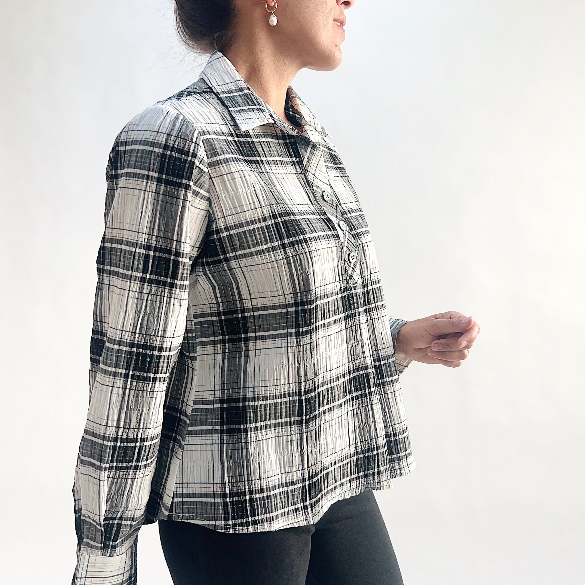 Side profile view of model wearing a white and charcoal windowpane plaid crinkle pullover shirt, showing the point collar, half placket, crinkle-texture fabric, and relaxed A-line silhouette, styled with black slim pants against a white background.
