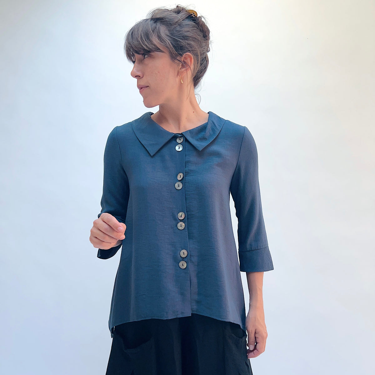 A woman wearing a slate blue button-up top with a pointed collar and three-quarter sleeves, paired with black pants, standing against a light gray background.
