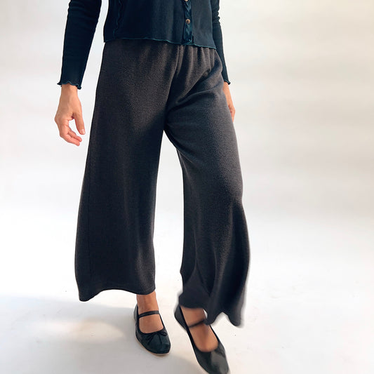 Model wearing the Cut Loose Marled Knit Crop Pant in Iron, close-up side view mid-stride, showing wide-leg silhouette, clean cropped hem, and dark charcoal marled knit texture, styled with a dark teal cardigan and black mary jane flats
