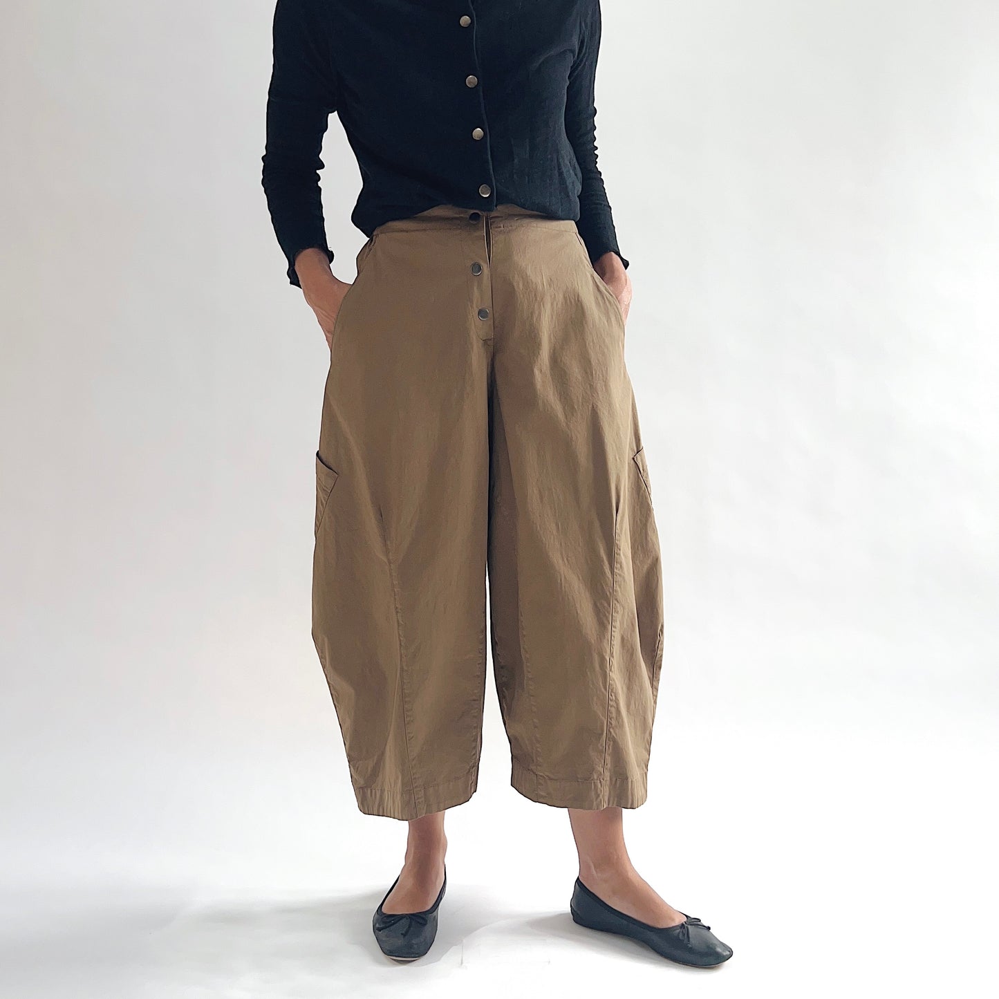 Full front view of model wearing Eleven Stitch winter olive snap front pant with hands in pockets, showing snap front waistband, wide balloon leg silhouette, large side patch pockets, gathered elastic ankle hem, and warm olive tan color, styled with black button front top and black ballet flats